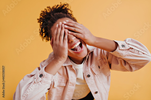 Excited black girl with ponytail covering eyes with hands. Studio shot of smilng joyful african woman in jacket. photo