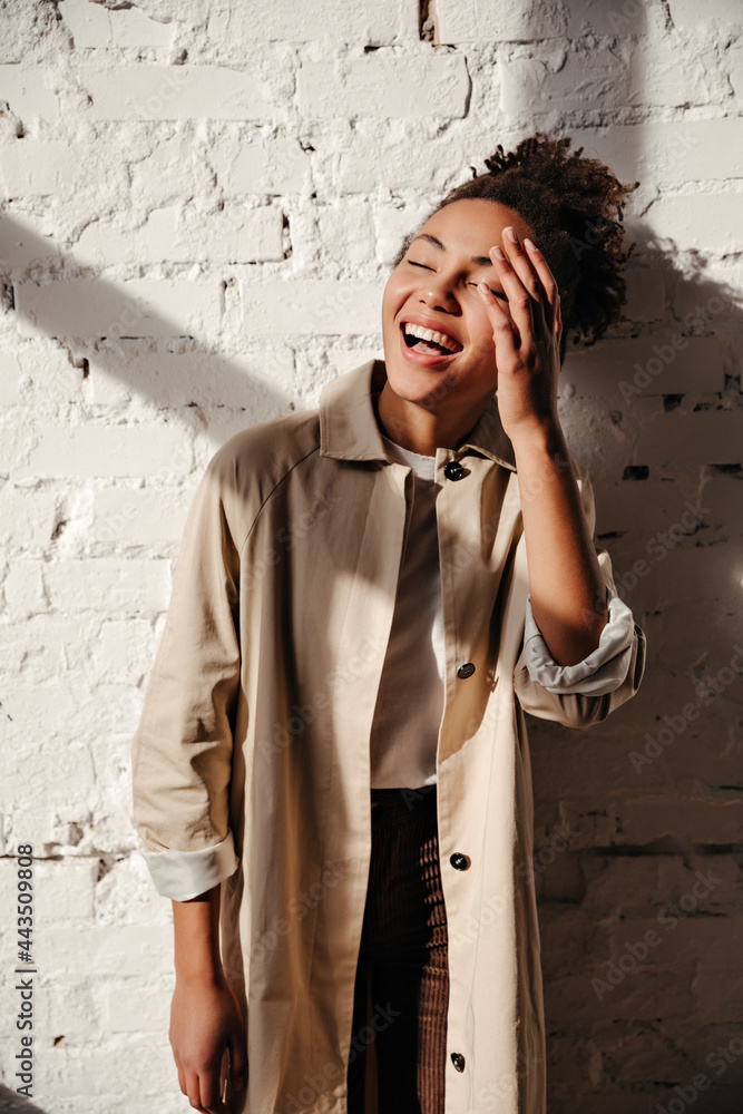Glamorous black woman in trench coat laughing with closed eyes. Indoor shot of wonderful african american girl standing near bricked wall under sun rays. Glamorous black woman in trench coat laughing with closed eyes. Indoor shot of wonderful african american girl standing near bricked wall under sun rays.