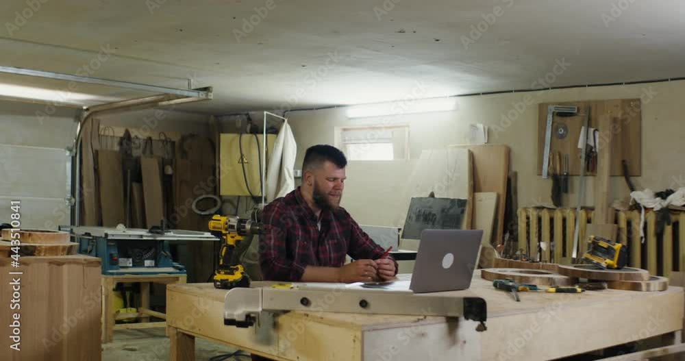 a european man talking by video call on a laptop in a carpentry workshop a european man talking by video call on a laptop in a carpentry workshop