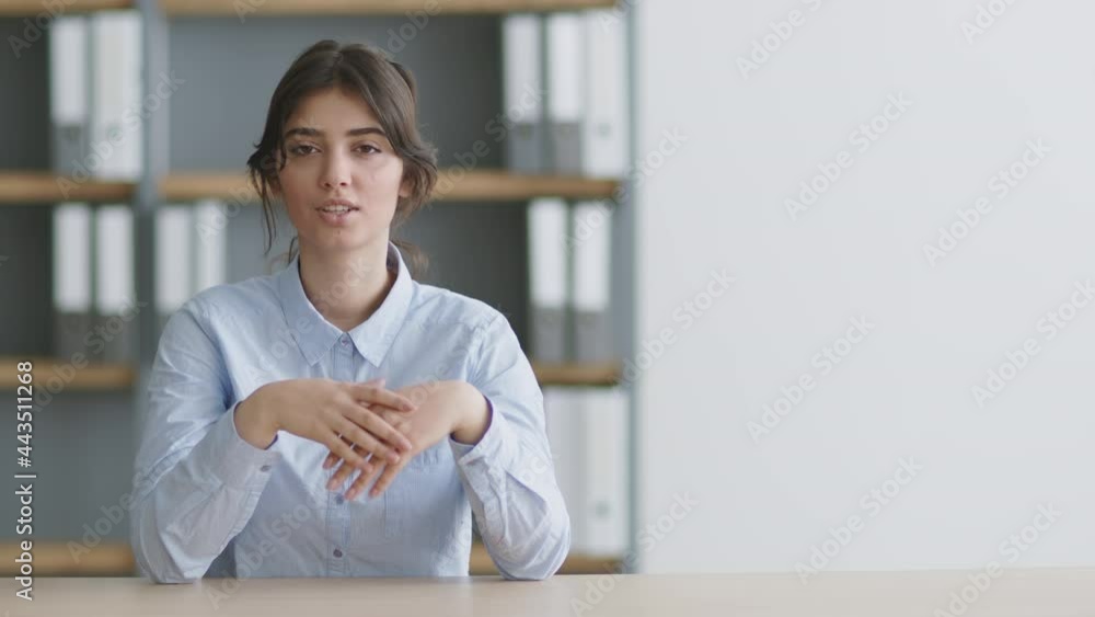 Young professional woman speaker talking to camera, explaining and pointing at empty space, sitting in office interior Young professional woman speaker talking to camera, explaining and pointing at empty space, sitting in office interior