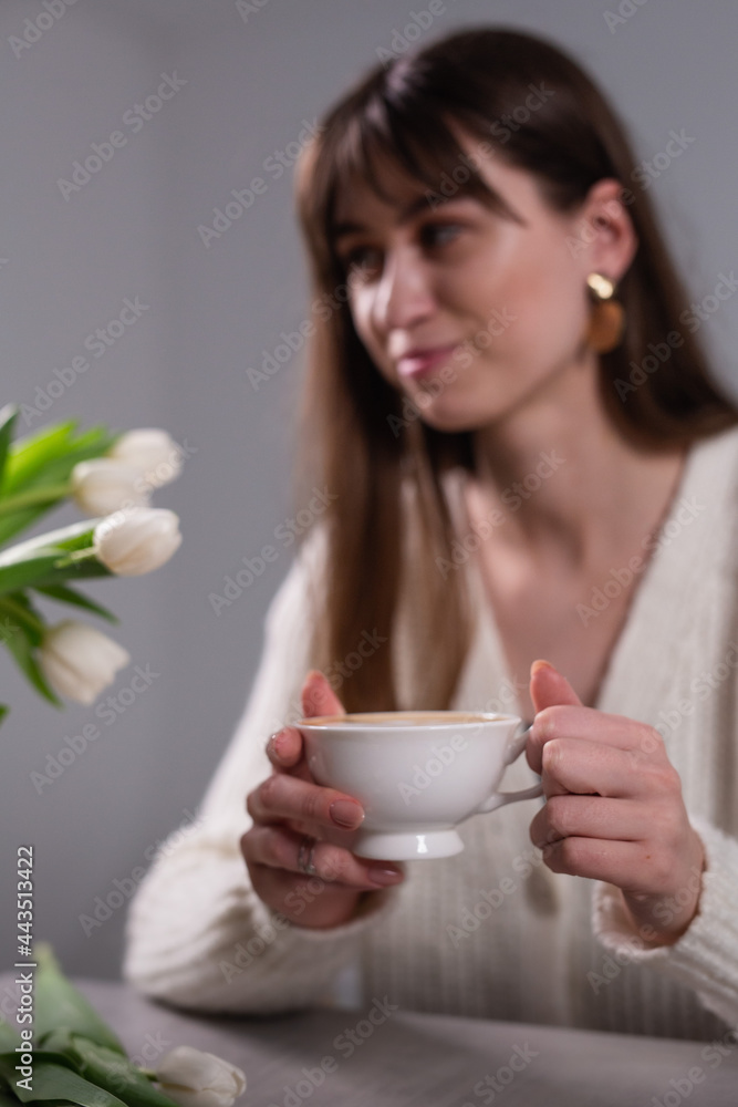 Beautiful girl with long hair and bangs holding a cup of cappuccino. A bouquet of tulips near the girl on the table. Beautiful girl with long hair and bangs holding a cup of cappuccino. A bouquet of tulips near the girl on the table.