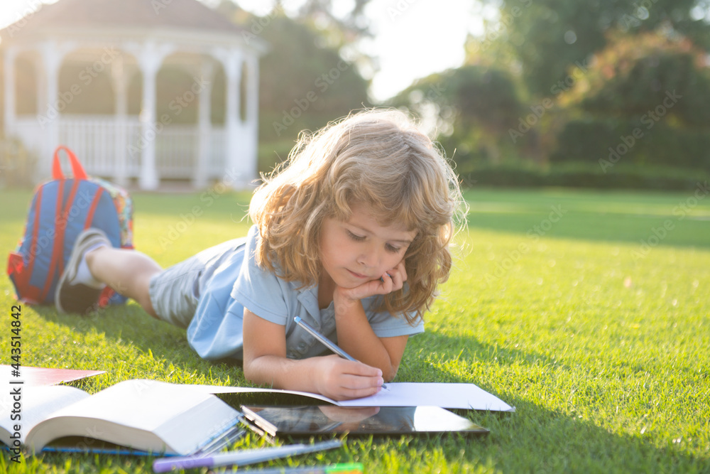 Beautiful child boy with book writing notes in copybook on grass background. Kids reading book in park. Beautiful child boy with book writing notes in copybook on grass background. Kids reading book in park.