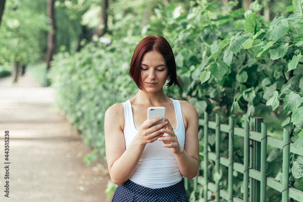 Beautiful red-haired girl pensively prints a message while walking in the park Beautiful red-haired girl pensively prints a message while walking in the park