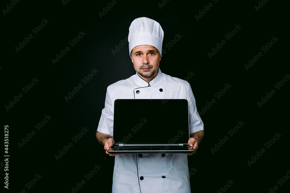 Chef cook wearing cooking jacket and hat, holding a laptop and showing its screen, on black background Chef cook wearing cooking jacket and hat, holding a laptop and showing its screen, on black background