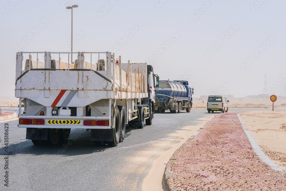 Group of lorry transports, delivery truck and tanker, on highway through the desert with blurred sand dunes on background due sand storm.