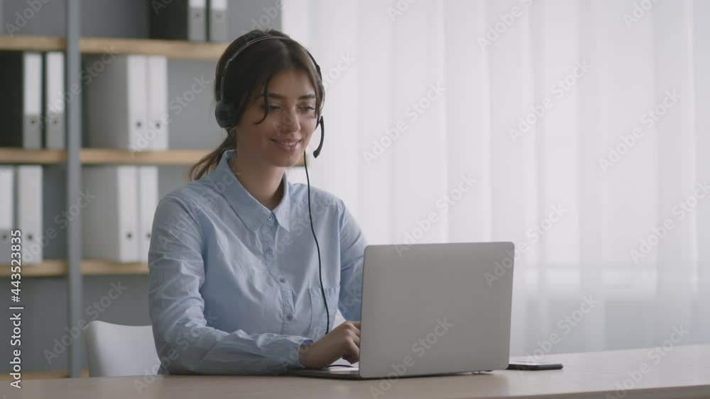 Online hotline center. Positive young woman operator in headset talking with customer, typing on laptop at office Online hotline center. Positive young woman operator in headset talking with customer, typing on laptop at office