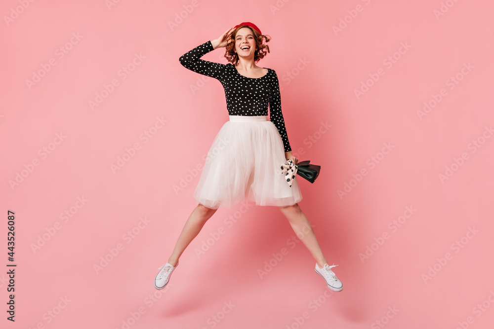 Pleased girl in white skirt jumping with smile. Full length view of curly woman having fun on pink background.