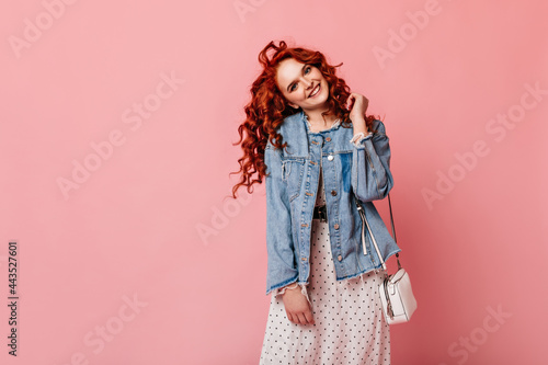 Playful ginger young woman looking at camera with smile. Studio shot of charming girl in denim jacket expressing happiness.