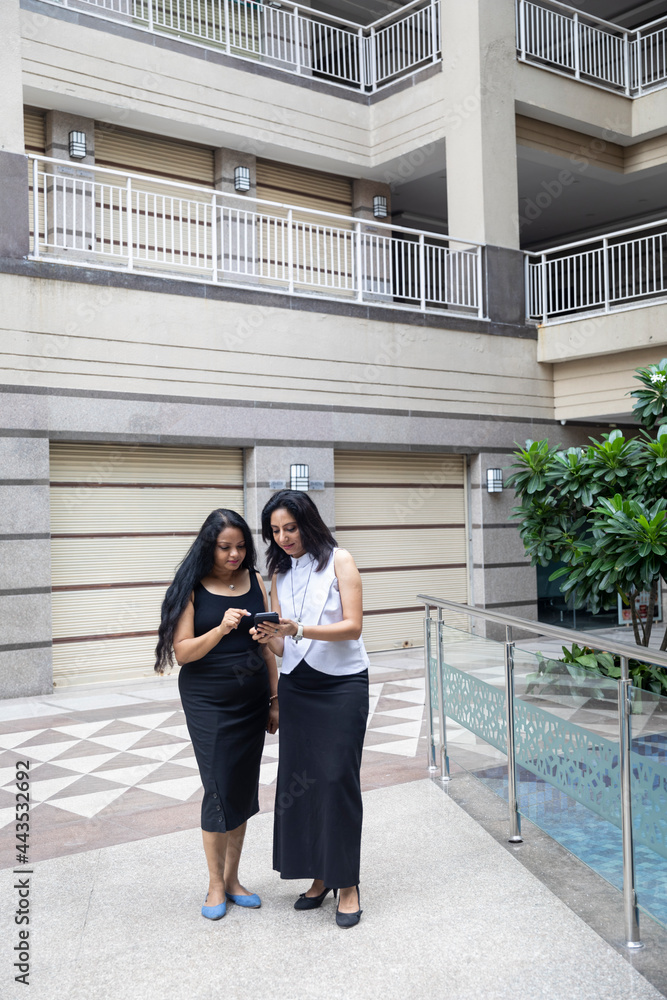 Two Indian woman having a discussion as they look into their phone in an urban corporate setting.