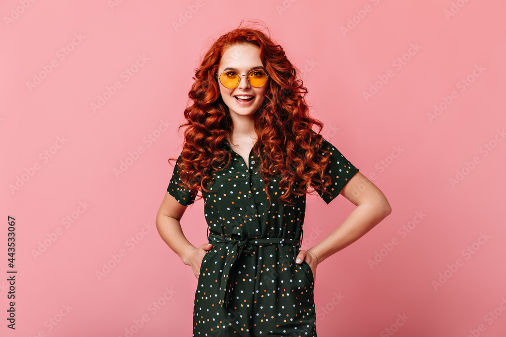 Appealing ginger girl standing with hands on hips. Happy curly young woman in sunglasses posing on pink background. Appealing ginger girl standing with hands on hips. Happy curly young woman in sunglasses posing on pink background.