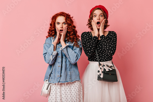 Amazed girl in denim jacket posing with friend. Two attractive women expressing surprised emotions on pink background.