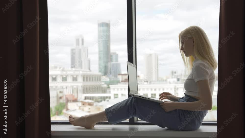 A young blonde woman works behind a laptop on the windowsill near a large window. A beautiful woman opens a laptop and starts typing on the keyboard. Freelance and remote work concept. 4K UHD A young blonde woman works behind a laptop on the windowsill near a large window. A beautiful woman opens a laptop and starts typing on the keyboard. Freelance and remote work concept. 4K UHD