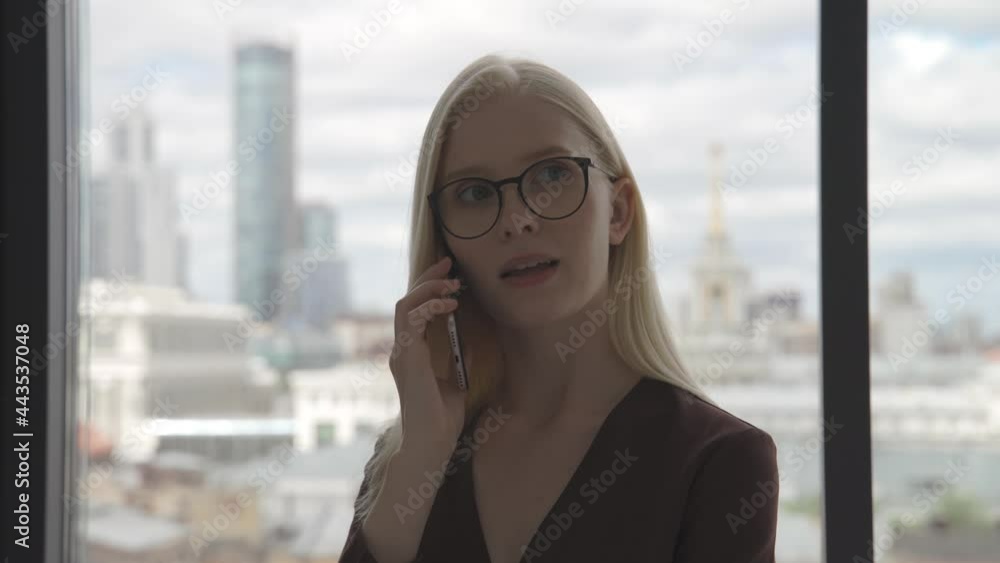 Young business woman talking on the phone near a large window overlooking the city center. Businesswoman in the office. Close-up, 4K UHD. Young business woman talking on the phone near a large window overlooking the city center. Businesswoman in the office. Close-up, 4K UHD.
