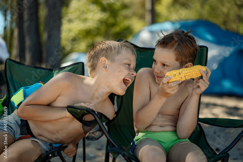 cute caucasian boys of elementrary age eating corn on beach sitting on camping chairs, laughing, enjoyig holiday photo