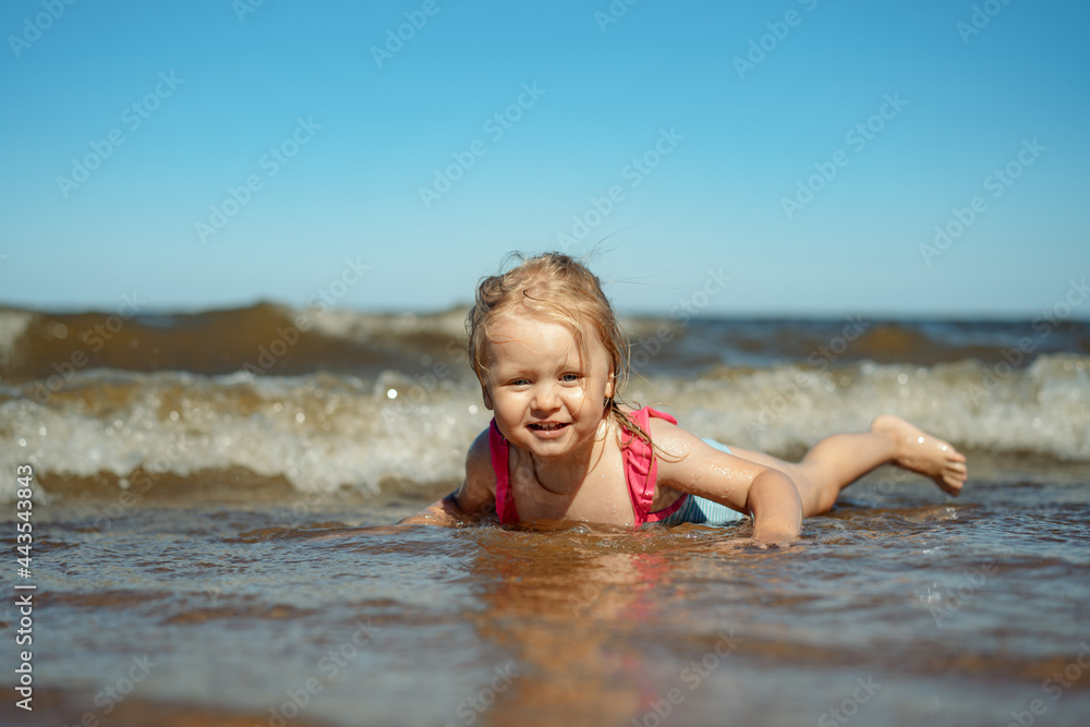 Cute Little caucasian blond girl lying in sea in waves. Vacation with kids. Sunny day on the beach Cute Little caucasian blond girl lying in sea in waves. Vacation with kids. Sunny day on the beach
