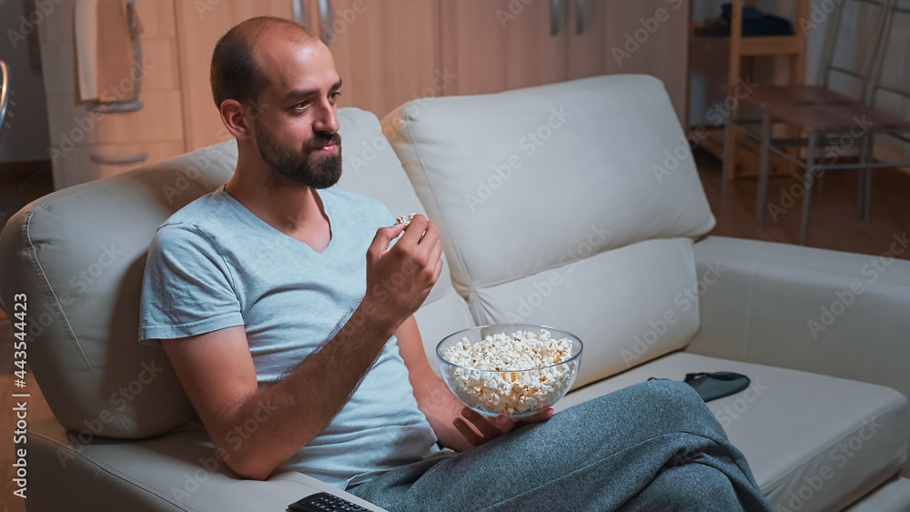 Caucasian male chilling on sofa with popcorn bowl in hands while watching movie series on television. Tired man in pajamas looking at entertainment shows late at night in kitchen Caucasian male chilling on sofa with popcorn bowl in hands while watching movie series on television. Tired man in pajamas looking at entertainment shows late at night in kitchen