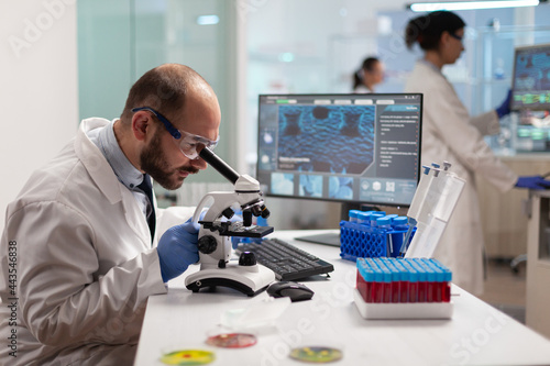 Microbiologist working to find a vaccine for virus in lab looking at sample through microscope weaing protection glasses. Specialists working on medicine, biotechnology research in advanced pharma lab photo