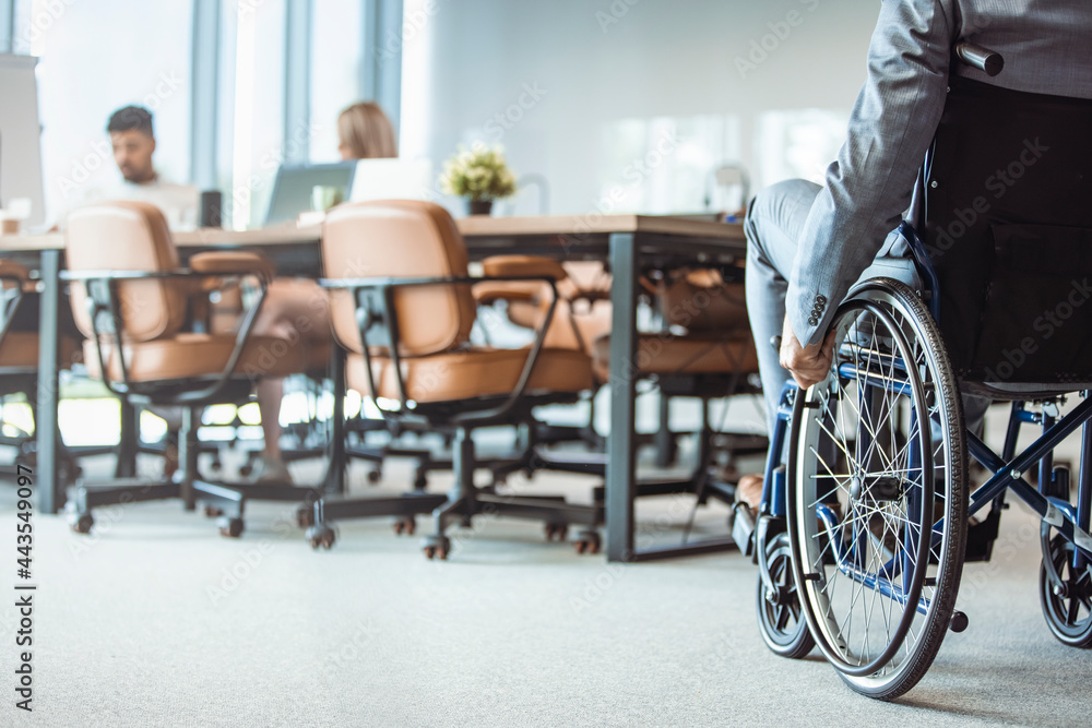Fototapeta premium Close up. Disabled Man on Wheelchair in Office. Disabled Young Man. Man on Wheelchair. Recovery and Healthcare Concepts. Teamwork in Office. Young Workers. Sitting Man. Happy Worker.