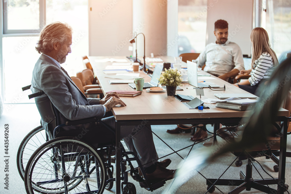 Portrait of sad disabled businessman sitting in wheelchair in office with colleagues working on background. Handicapped lonely male employee in wheelchair sitting in office Portrait of sad disabled businessman sitting in wheelchair in office with colleagues working on background. Handicapped lonely male employee in wheelchair sitting in office