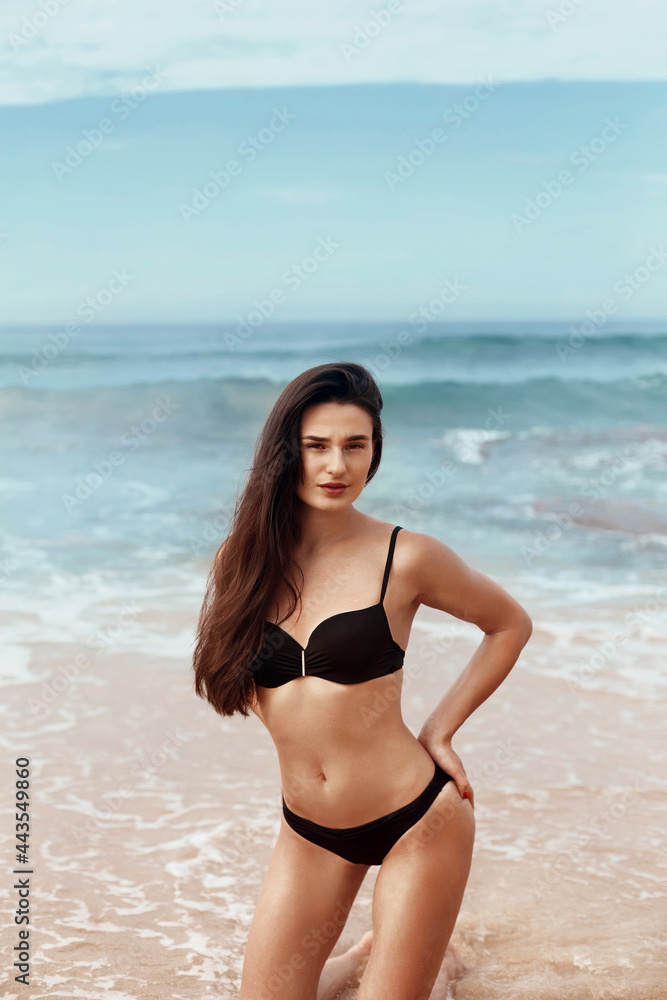 Portrait of young woman in black bikini on tropical beach looking at camera. Beautiful  girl in swimwear with copy space. Summer vacation and tanning concept.