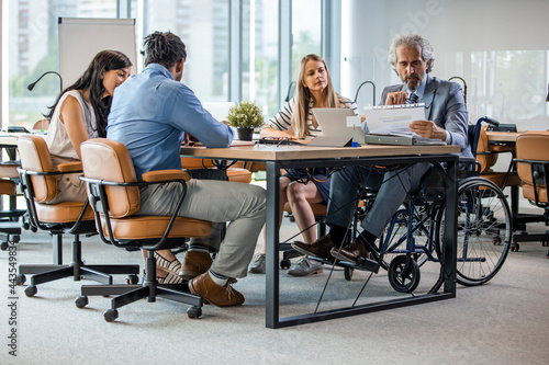 Happy businessman in wheelchair reading documents during a meeting with his colleagues in the office. Shot of a team of businesspeople having a meeting in a modern office