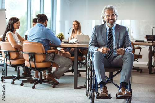 Fototapeta Naklejka Na Ścianę i Meble -  Confident and modern architect in a wheelchair, working in his office. Wheelchair businessman portrait at workplace. They're a diverse and dynamic team. Diverse Group of Colleagues