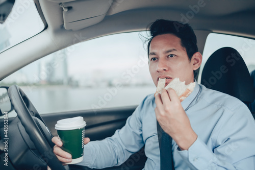 A businessman eating whole wheat toast and holding a coffee cup While driving on the road and he did not lose a seat belt.