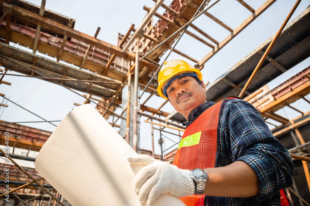Worker man in hard hat holding blueprint and looking at camera checking and planning project at the construction site Worker man in hard hat holding blueprint and looking at camera checking and planning project at the construction site