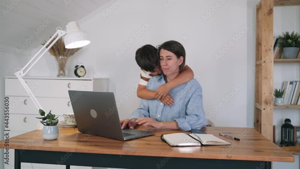 A woman with a child is working at a laptop. The boy hugs and kisses his mother while working. A woman kisses her son. Support in the family. Freelance. Self-isolation. Work at home A woman with a child is working at a laptop. The boy hugs and kisses his mother while working. A woman kisses her son. Support in the family. Freelance. Self-isolation. Work at home