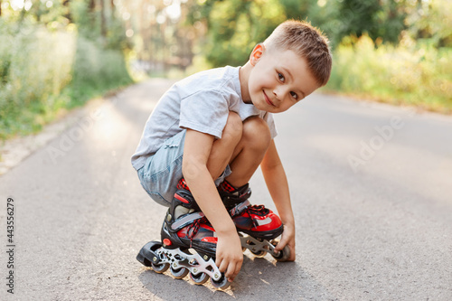 Boy in roller skating sitting on the road, looking at camera with smile, wearing white t-shirt and short, child squats while having rest rollerblading.