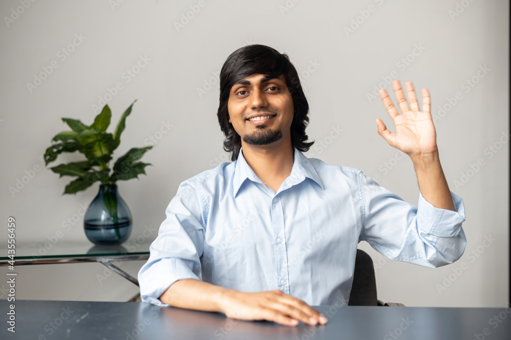 Nice to see you.Cheerful indian colleague waving hand into the camera, happy hindu guy in formal shirt sits at the desk and greeting online interlocutor, takes a part in online conference,webcam view Nice to see you.Cheerful indian colleague waving hand into the camera, happy hindu guy in formal shirt sits at the desk and greeting online interlocutor, takes a part in online conference,webcam view