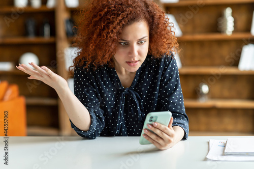 Shocked red-haired curly woman holding a smartphone, feel astonishment with a bad news, fired from work, irritated female eoffice employee looks at phone screen and does not understand what happened