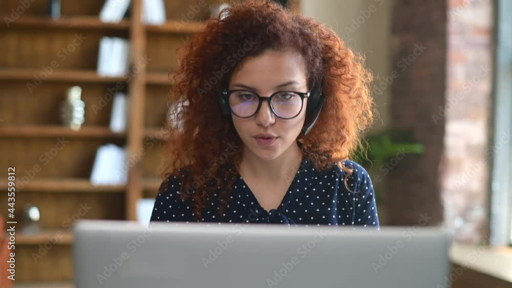 Confident red-haired curly woman wearing headset looks at the webcam of the laptop and talking online, female employee chatting with colleagues or customer on the distance. Video call, virtual meeting Confident red-haired curly woman wearing headset looks at the webcam of the laptop and talking online, female employee chatting with colleagues or customer on the distance. Video call, virtual meeting
