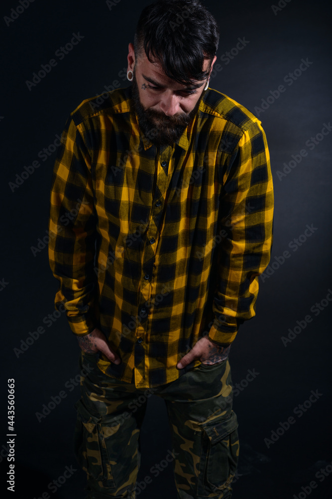 Male model with tattoos poses with hands in his pocket wearing yellow and black checkered shirt and army pants
