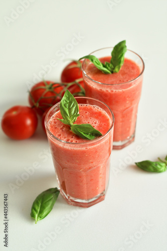 Two glasses of fresh organic tomato juice decorated with raw tomatoes and green leaves of basil on light background.Healthy diet nutrition concept.