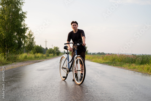 Young woman standing alone on road with bicycle looking at camera at countryside. Summer activity, healthy lifestyle, workout, sport, fitness. Person in nature. Riding bicycle. Having fun outdoors.