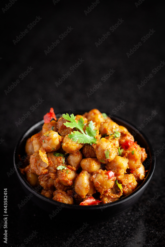 Chickpeas masala (Spicy chola or chhole curry) garnished with fresh green coriander and ingredients. Served in a black bowl. A Classic Indian typical Panjabi street food. Chickpeas masala (Spicy chola or chhole curry) garnished with fresh green coriander and ingredients. Served in a black bowl. A Classic Indian typical Panjabi street food.