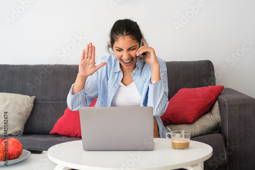 Young excited latin casual woman on mobile phone call using laptop at home. Expressive girl gesturing. photo