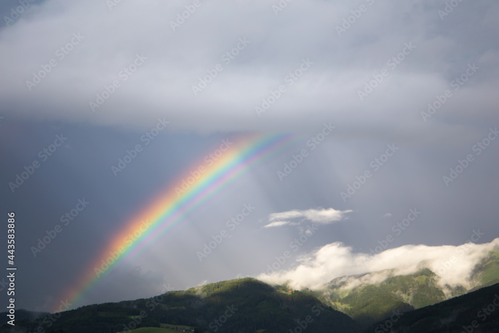 Fototapeta premium Rainbow on a dark grey sky background with some rays of sun shining through the clouds. On the bottom of the picture are some hills behind white clouds.