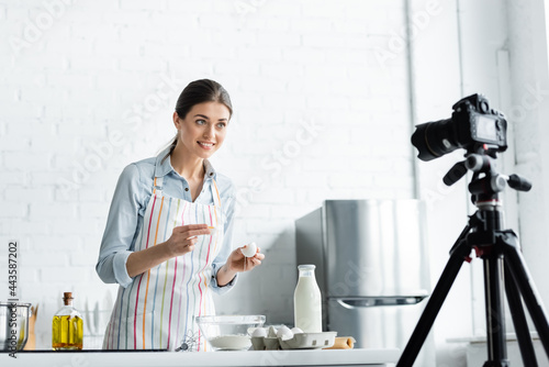 happy culinary blogger looking at blurred digital camera while breaking chicken egg into bowl with flour. photo