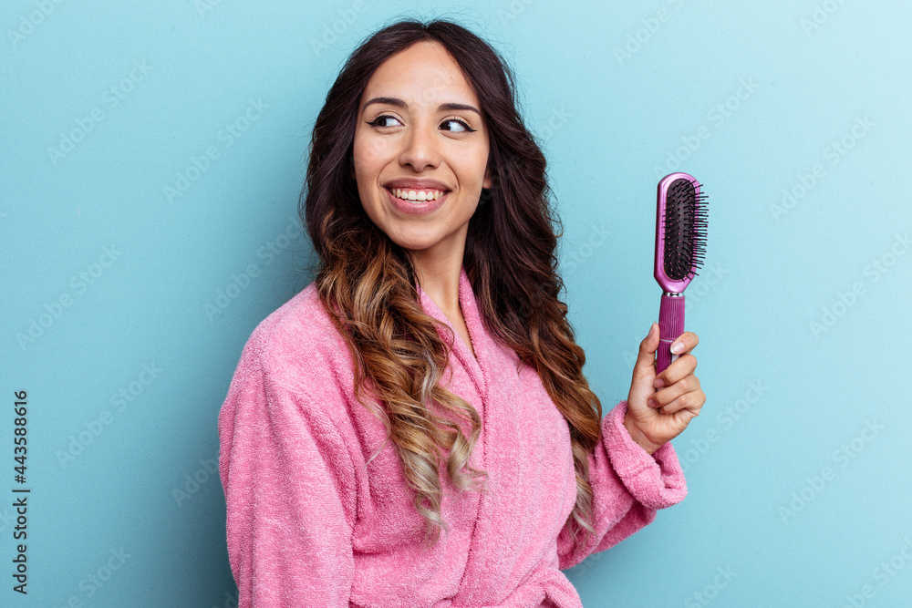 Young mexican woman wearing a bathrobe holding a brush isolated on blue background looks aside smiling, cheerful and pleasant. Young mexican woman wearing a bathrobe holding a brush isolated on blue background looks aside smiling, cheerful and pleasant.