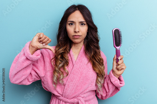 Young mexican woman wearing a bathrobe holding a brush isolated on blue background showing a dislike gesture, thumbs down. Disagreement concept.