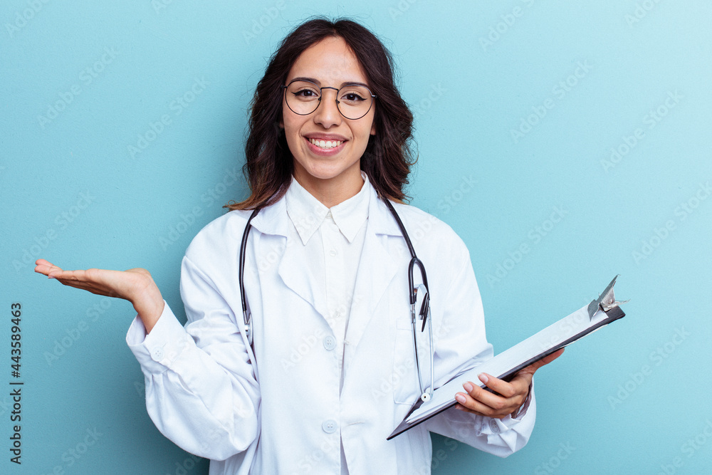 Young doctor mexican woman isolated on blue background showing a copy space on a palm and holding another hand on waist.
