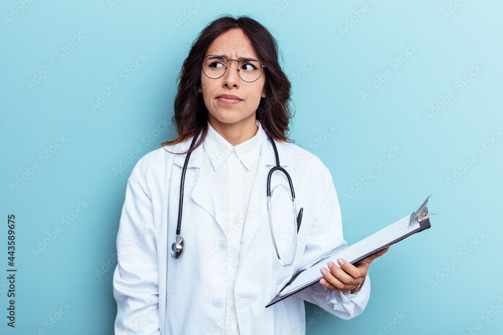Young doctor mexican woman isolated on blue background confused, feels doubtful and unsure. Young doctor mexican woman isolated on blue background confused, feels doubtful and unsure.