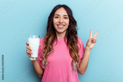 Young mixed race woman holding a glass of milk isolated on blue background joyful and carefree showing a peace symbol with fingers.