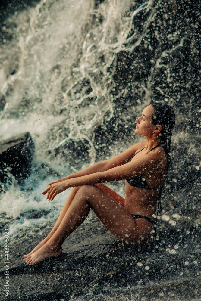 Fototapeta premium Beautiful woman sits under the waterfall and enjoys the falling water.
