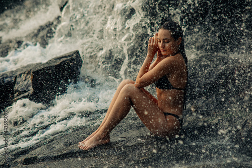 Beautiful woman sits under the waterfall and enjoys the falling water.