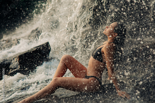 Beautiful woman sits under the waterfall and enjoys the falling water.