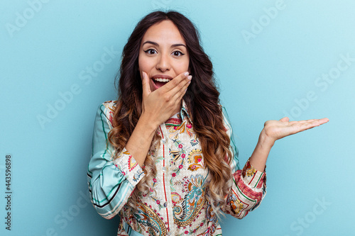 Young mexican woman isolated on blue background holds copy space on a palm, keep hand over cheek. Amazed and delighted.