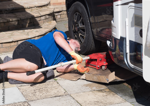 A male mechanic lying down as he places a red trolley jack under front of a motorhome.Recreational vehicle is on a private driveway